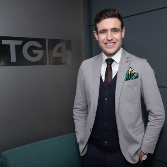 A half body shot of the man wearing a grey suit and smiling with hands in pockets standing to the right. The background has a TG$ sign on the left.