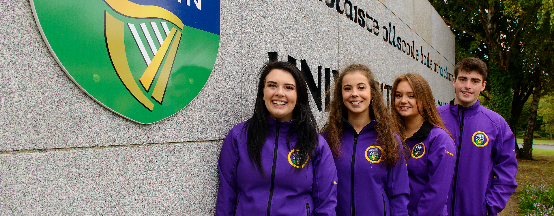 Gaeltacht UCD students standing beside the UCD logo