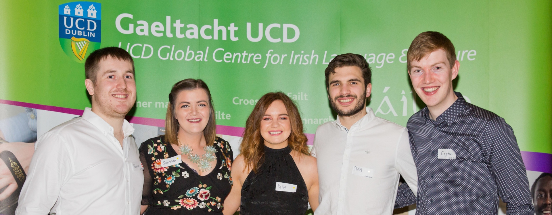 Five Gaeltacht UCD students standing and smiling together in front of the Gaeltacht UCD banner filling the background