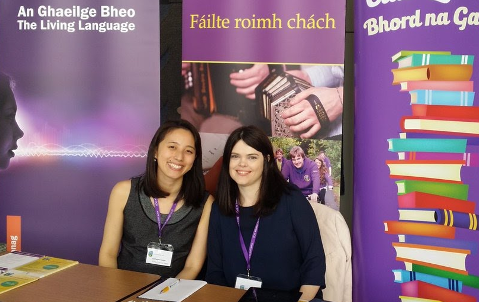Two Irish speaking employees sitting in front of a desk with failte roimh chach banners in the background