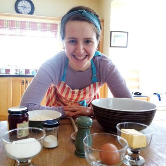 A kitchen table with bakery ingredients with a smiling woman leaning on it with a kitchen in the background