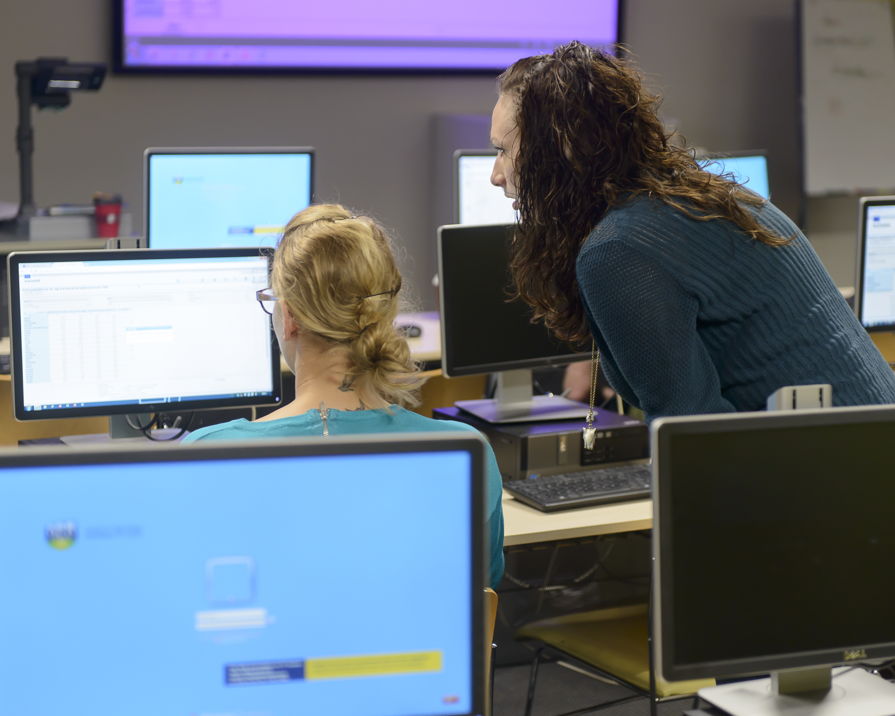 Photo of a student sitting at a computer screen with another person looking at the screen over her shoulder, presumably to help