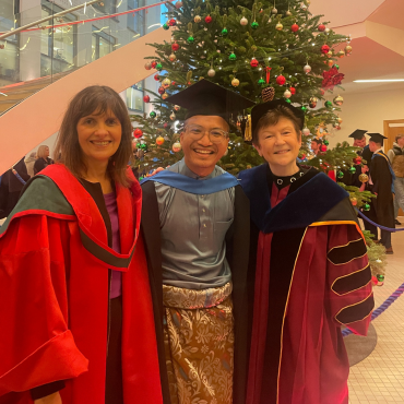 Graduate in cap with two female academics beside Christmas tree