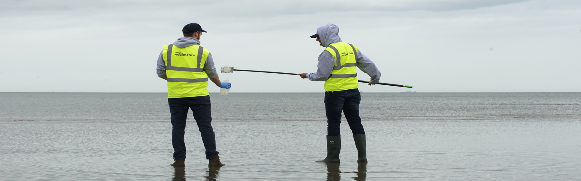 Two researchers at the beach wearing high-vis collecting water samples.