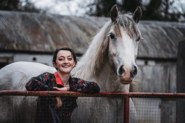 Orla Byrne leaning on a gate with a grey horse standing beside her